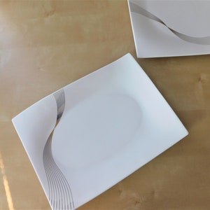 Peut inclure: Deux assiettes rectangulaires blanches avec un motif ondulé argenté. Les assiettes sont sur une surface en bois brun clair.