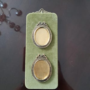 May include: A rectangular olive green velvet display board featuring two oval-shaped metallic picture frames. Each frame has an ornate, textured border and a small decorative bow detail at the top. The frames are mounted vertically on the velvet.