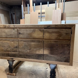 Peut inclure: Un grand buffet en bois rustique avec six tiroirs. Le meuble est fait de bois récupéré avec une finition naturelle et vieillie. Il est soutenu par des pieds sombres et sculptés et se trouve dans un atelier.