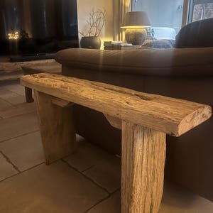 Könnte beinhalten: Rustikale Holzbank mit natürlicher, verwitterter Optik. Die Bank besteht aus dicken, rechteckigen Holzbohlen, die Maserung und Textur zeigen. Die Beine sind ebenfalls aus Holz und bieten eine stabile Basis. Geeignet für den Innenbereich.