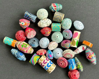 May include: A collection of colorful paper beads in various shapes and sizes. The beads feature a variety of patterns and colors, including red, blue, green, and gold. Some beads are cylindrical, while others are round or oval.