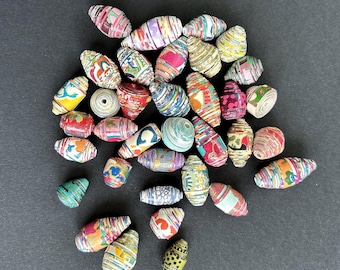 May include: A collection of colorful paper beads in various shapes and sizes. The beads feature vibrant patterns and designs in shades of red, blue, yellow, and green. Some beads are cylindrical, while others are oval or round.