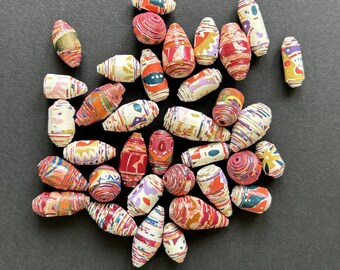 May include: A collection of colorful paper beads in various shapes and sizes. The beads feature a mix of red, orange, yellow, and blue patterns. Some beads have a spiral design, while others have geometric shapes. The beads are scattered on a dark surface.
