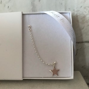 May include: A silver earring with a star pendant and chain, presented in a white gift box. The earring features a small ball stud, a delicate chain, and a five-pointed star charm. A white ribbon with text is visible.