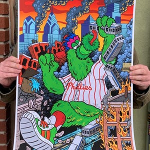May include: A colorful art print featuring a green, furry mascot in a Phillies baseball uniform, holding a train. The background depicts a city with burning buildings and smoke, with the word "Phillies" on the jersey.