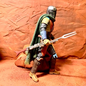 Puede incluir: Una figura de acci&oacute;n de un guerrero mandaloriano, con un casco y una armadura verde y marr&oacute;n, sosteniendo una lanza plateada y blanca. La figura est&aacute; de pie sobre una roca.