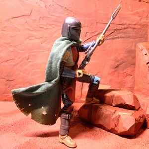 Puede incluir: Una figura de acci&oacute;n de un guerrero mandaloriano con una capa verde y sosteniendo un arma similar a una lanza. La figura est&aacute; de pie sobre una superficie rocosa, con un fondo rojo-marr&oacute;n.
