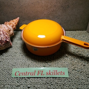 May include: A bright yellow enameled metal skillet with a matching lid and a long handle. A large, textured, tan and brown seashell sits to the left. A rectangular green sign with the red text Central FL skillets is placed in the foreground.