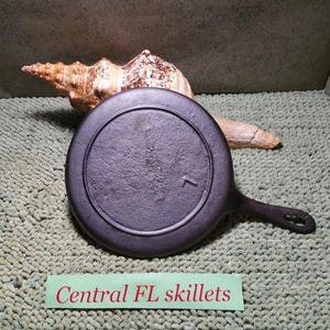 May include: A black cast iron skillet with a handle, resting on a textured surface. A seashell and a piece of driftwood are in the background. The text "Central FL skillets" is on a green label.