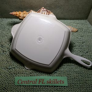 May include: A white square skillet with a handle, resting on a green textured surface. The skillet has a raised edge and a circular marking in the center. The text "Central FL skillets" is visible.