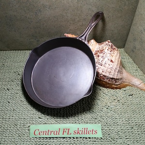 May include: A dark gray cast iron skillet with a long handle. The skillet is round with a slightly curved edge. The text "Central FL skillets" is visible on a green label. A large seashell is in the background.