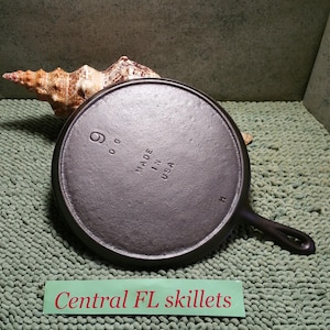 May include: A black cast iron skillet with a handle, marked "9" and "Made in USA." The skillet is on a textured green surface, with a seashell in the background. The text "Central FL skillets" is on a green label.