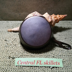 May include: A dark, cast iron skillet with a single handle, resting on a textured, light green surface. A large seashell is behind the skillet. The text "Central FL skillets" is on a green label.