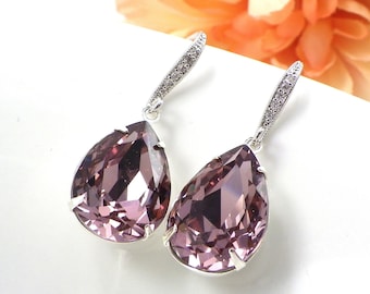 Puede incluir: Un par de elegantes pendientes colgantes con grandes cristales en forma de pera de color lavanda. Los pendientes tienen un engaste plateado y una sección superior delgada y brillante. El fondo incluye una superficie blanca y una flor naranja suave.