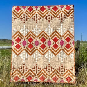 May include: A large, rectangular quilt with a geometric pattern of red, orange, pink, and white diamonds and chevrons on a beige background. The quilt is hanging outdoors, with a blue sky and green field in the background.
