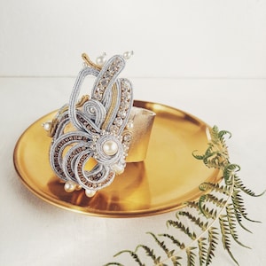 Puede incluir: Un brazalete plateado y dorado con un diseño de cuentas y detalles de perlas. El brazalete está en una bandeja dorada con una hoja de helecho verde.