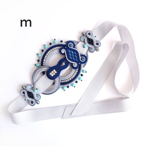 Puede incluir: Una cinta plateada con un diseño trenzado y con cuentas azules y turquesas. El diseño presenta un círculo con un círculo más pequeño en su interior. La cinta está atada en un lazo.