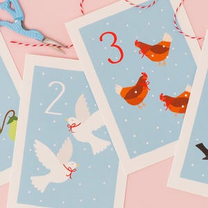 May include: Four illustrated cards with numbers 1-4, featuring birds and fruit on a light blue background. The cards are decorated with a partridge, doves, hens, and blackbirds. A pair of blue scissors and red and white twine are in the background.