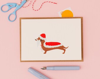 May include: A greeting card featuring a brown dachshund wearing a Santa hat and red coat with white trim. The card is on a brown border. A light blue pen and scissors, along with red and white twine, are on a pink background.