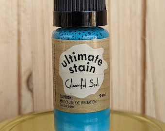 Può includere: Un piccolo flacone di vernice blu etichettato "Ultimate Stain Colourful Soul" con un tappo nero. Il flacone è da 9 ml e ha un'etichetta di avvertenza che dice "Attenzione: Può causare irritazione agli occhi. Vedi il pannello laterale."