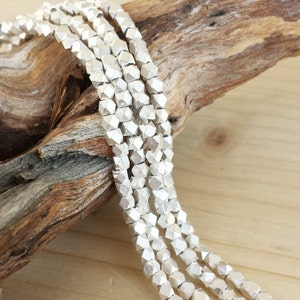 May include: Strands of silver-colored, geometric beads. The beads have a faceted, cube-like shape and are strung together. The beads are displayed on a piece of driftwood against a light wood background. The beads are likely for jewelry making.