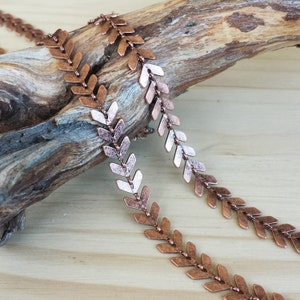 May include: A copper-colored chain with arrowhead-shaped links. The chain is lying on a piece of driftwood.