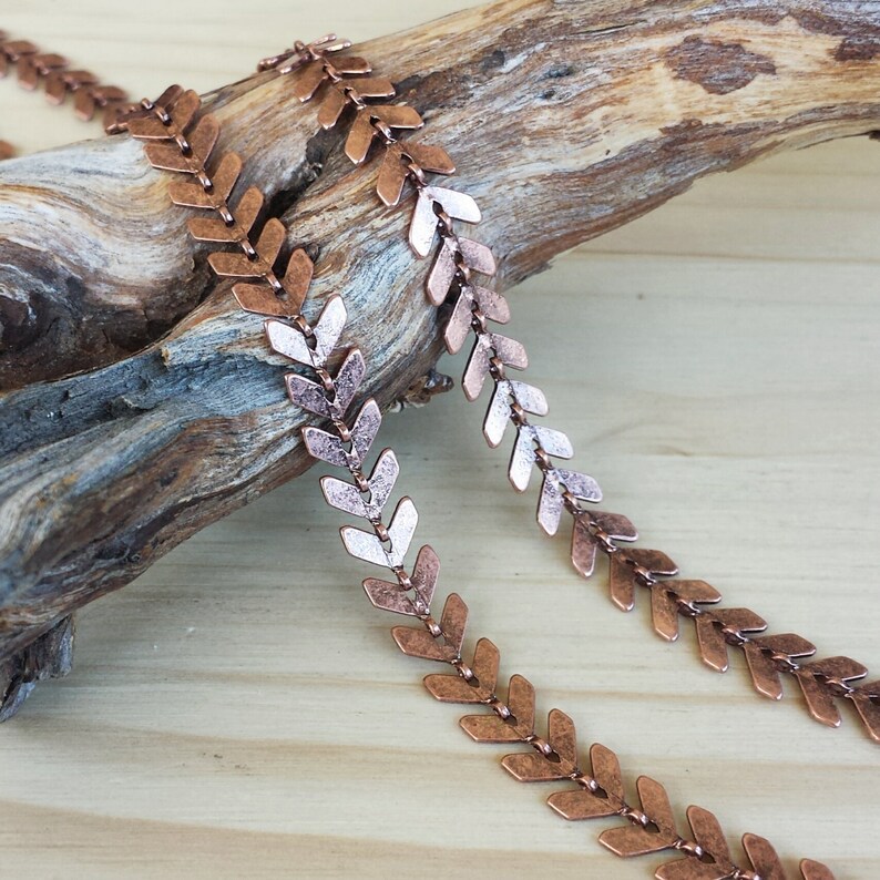 May include: A copper-colored chain with arrowhead-shaped links. The chain is lying on a piece of driftwood.
