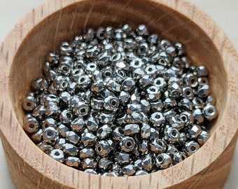 Op de afbeelding: Een houten kom gevuld met talrijke kleine, facetgeslepen, metalen kralen. De kralen hebben een glanzende, zilvergrijze uitstraling en zijn dicht op elkaar gepakt. De kom is gemaakt van lichtgekleurd hout, wat een contrast creëert met de kralen.