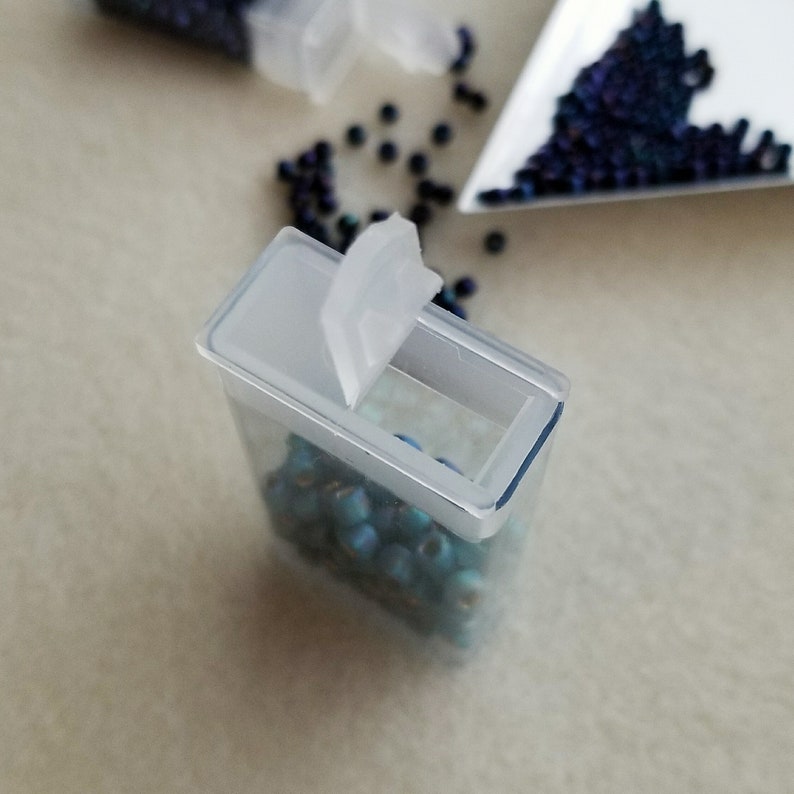 May include: A clear plastic container with a hinged lid, filled with small turquoise blue beads. The container is tilted slightly, revealing the beads inside.