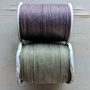 May include: Two spools of cord, one above the other. The top spool is a dark purple-brown color, and the bottom spool is a muted olive green. The cord is tightly wound on white cardboard spools, ideal for crafting or jewelry making.