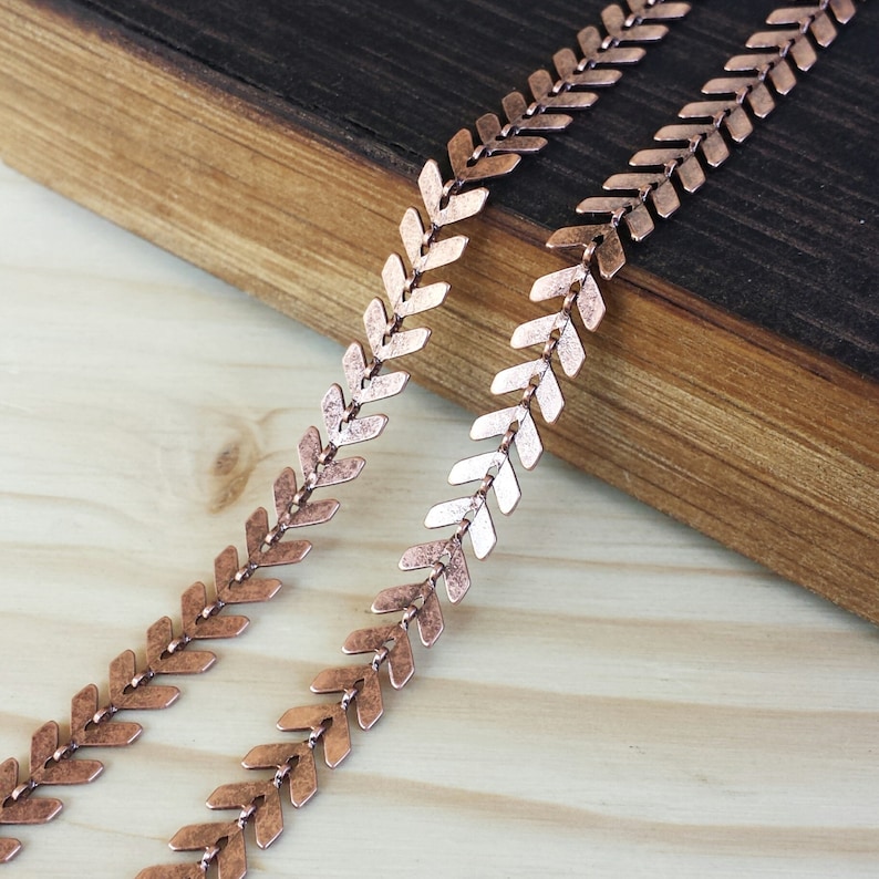 May include: Two strands of copper-colored chain with arrowhead-shaped links. The chain is lying on a wooden surface.