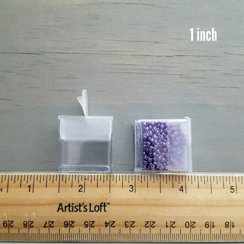 May include: Two clear plastic containers, one with a hinged lid, measuring 1 inch in height. The container on the right is filled with small, light purple beads.