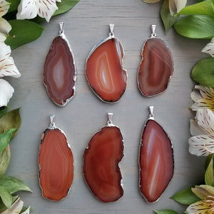 Puede incluir: Seis colgantes de rodajas de ágata con engarces de plata. Las rodajas de ágata son todas de diferentes formas y tamaños, pero todas tienen un esquema de color naranja y marrón similar.