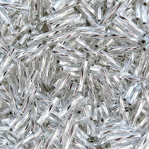 May include: A close-up shot displays a multitude of shiny, silver-colored glass beads. The beads are cylindrical with a slightly faceted surface, reflecting light. They are scattered, creating a textured, shimmering effect. Ideal for jewelry making and crafts.