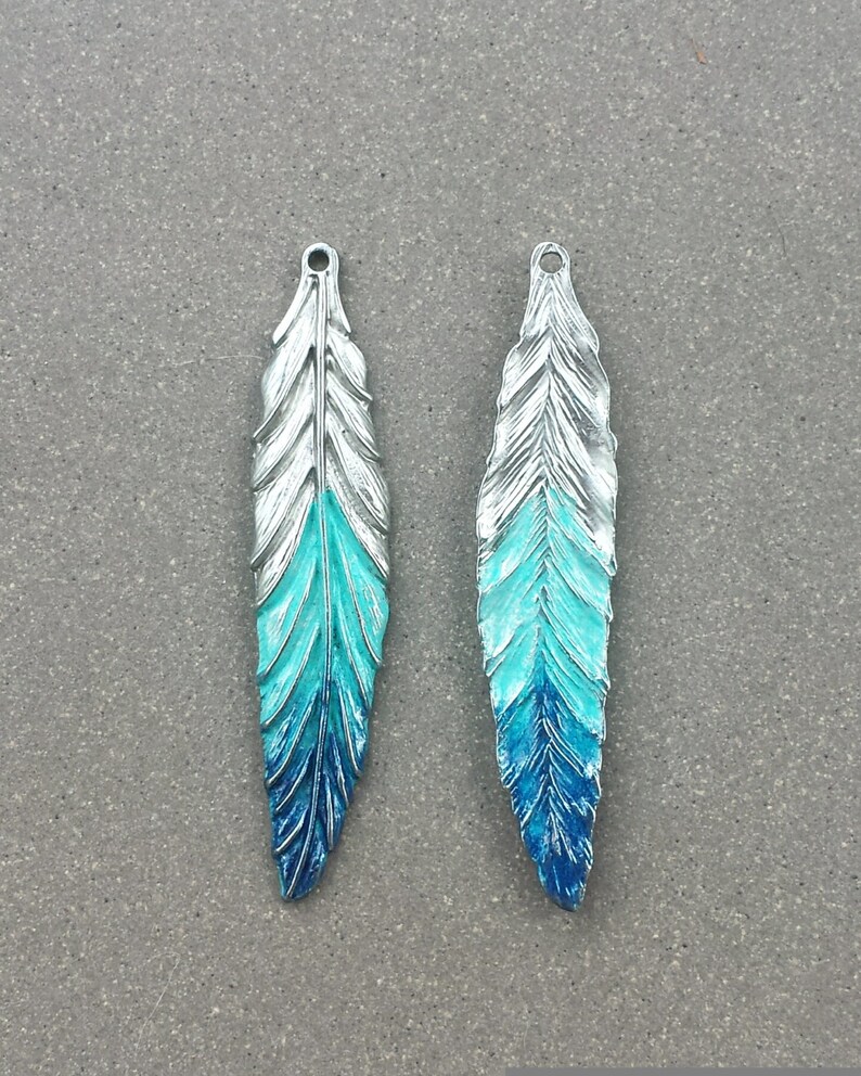 May include: Two silver-toned metal feather charms with a blue and turquoise ombre paint finish.