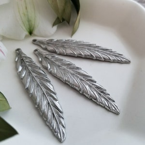 Op de afbeelding: Drie zilveren metalen veercharms met een gedetailleerd, gestructureerd ontwerp. De veren zijn in een rij op een wit oppervlak gerangschikt.