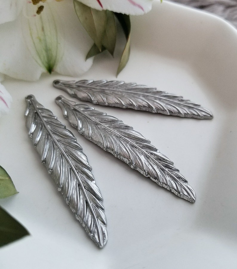 Op de afbeelding: Drie zilveren metalen veercharms met een gedetailleerd, gestructureerd ontwerp. De veren zijn in een rij op een wit oppervlak gerangschikt.