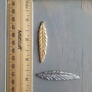 Op de afbeelding: Twee metalen verencharms, een gouden en een zilveren, met een liniaal als referentie voor de grootte. De gouden verencharm is ongeveer 5 cm lang. De zilveren verencharm is ongeveer 4 cm lang.