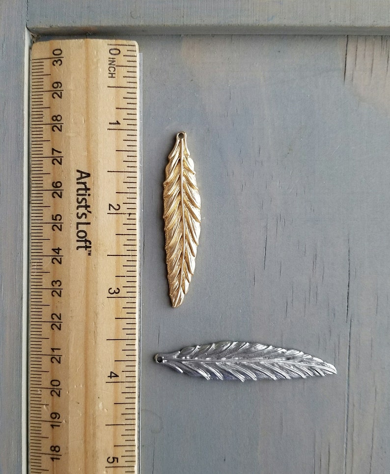 Op de afbeelding: Twee metalen verencharms, een gouden en een zilveren, met een liniaal als referentie voor de grootte. De gouden verencharm is ongeveer 5 cm lang. De zilveren verencharm is ongeveer 4 cm lang.