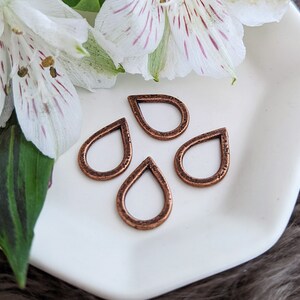 May include: Four copper-colored teardrop-shaped charms arranged on a white ceramic dish. The charms have a textured surface and are open in the center. White and purple-striped flowers are in the background.