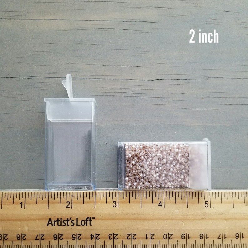 May include: A clear plastic container with a hinged lid, measuring 2 inches in height, next to a smaller container filled with small, white beads. A ruler is placed below the containers, showing the measurement of 2 inches.