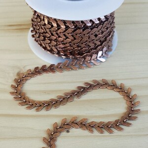 May include: A spool of copper-colored chain with a chevron pattern. The chain is made of small, flat links that are connected to form a continuous loop. The chain is wound around a white spool.
