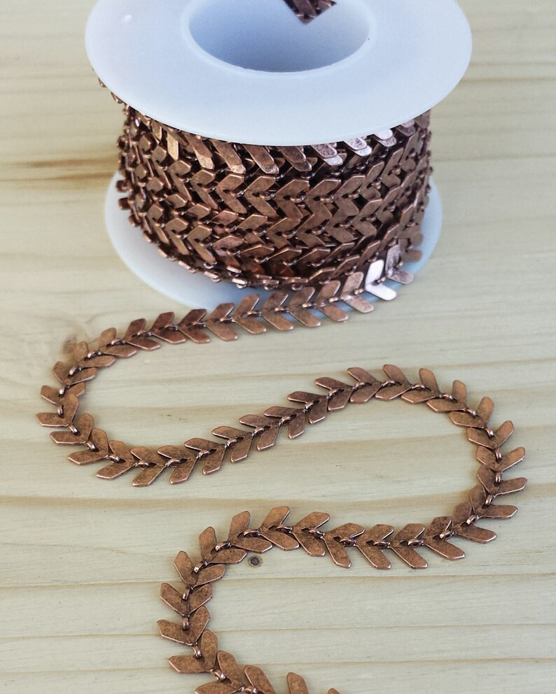 May include: A spool of copper-colored chain with a chevron pattern. The chain is made of small, flat links that are connected to form a continuous loop. The chain is wound around a white spool.