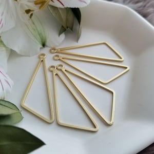 May include: Several gold-colored geometric earring components are arranged on a white surface. The components are open-frame shapes, including triangles and diamond shapes, each with a small loop at the top for attachment. The background includes white flowers and greenery.