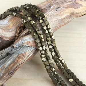 May include: A strand of gold-toned metal beads with a faceted, geometric shape. The beads are strung on a wire and are arranged in a row. The beads are lying on a piece of driftwood.