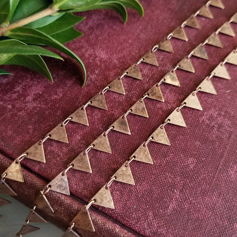 May include: Three strands of copper-toned chain with triangle-shaped links. The chain is lying on a burgundy fabric surface.