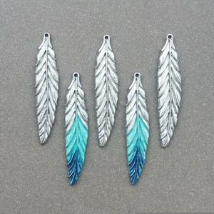 Op de afbeelding: Vijf zilveren verencharms met een blauwe en turquoise ombre-verflaag. De charms hebben een gaatje bovenaan om aan sieraden of andere knutselwerkjes te bevestigen.