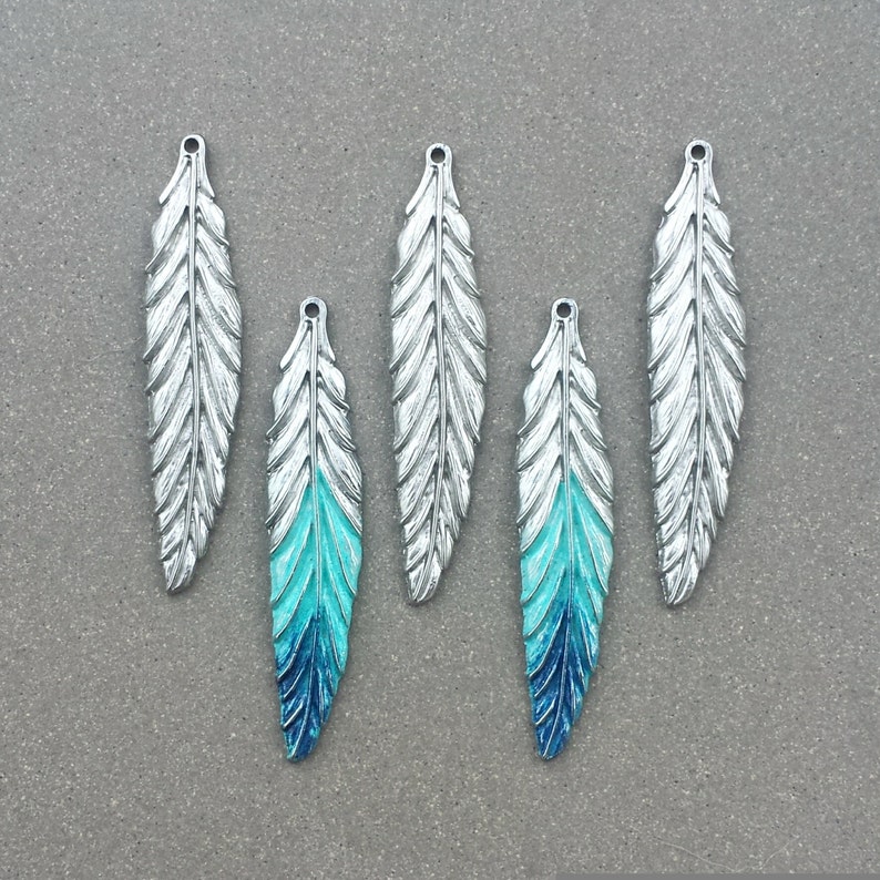 Op de afbeelding: Vijf zilveren verencharms met een blauwe en turquoise ombre-verflaag. De charms hebben een gaatje bovenaan om aan sieraden of andere knutselwerkjes te bevestigen.