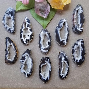 May include: A selection of polished agate slices in black, white, and grey. Each slice features a hollow centre with a crystalline interior. The agate pieces are arranged on a neutral surface, with crystals and flowers in the background.