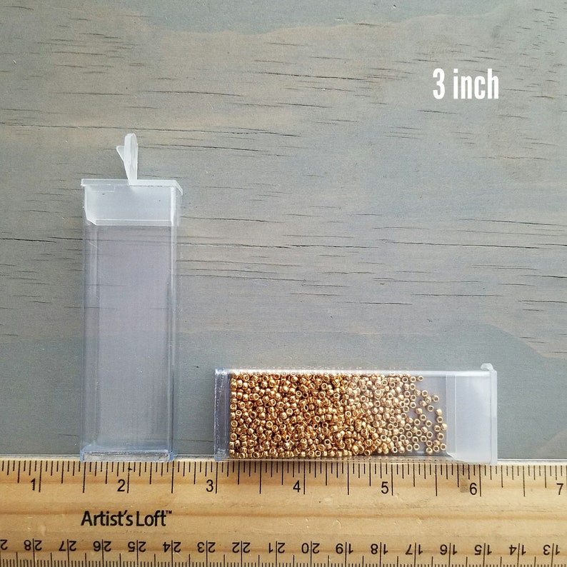 May include: Two clear plastic containers filled with gold seed beads. The containers are 3 inches long and are sitting on a wooden ruler. The ruler is marked with inches and fractions of an inch.